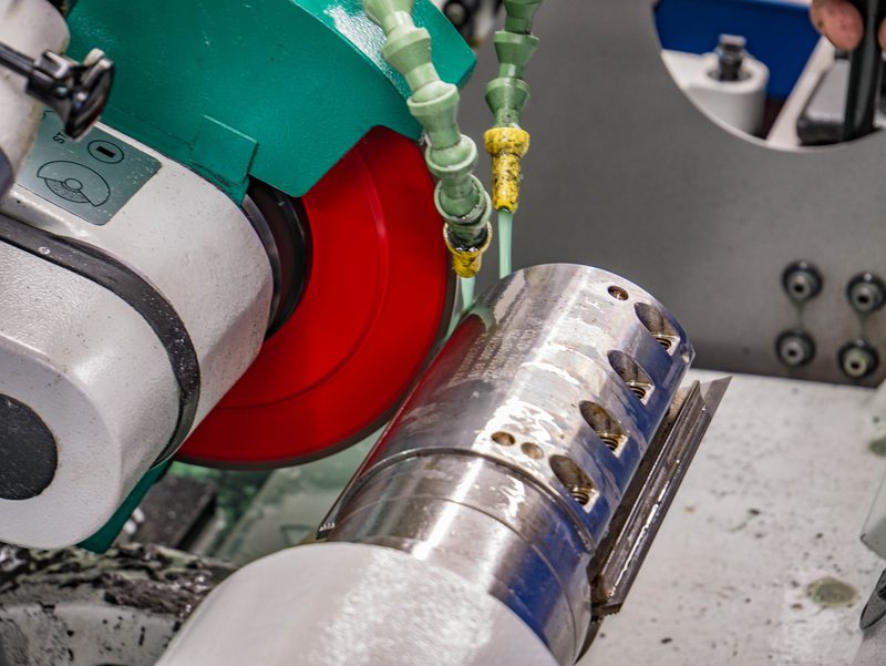 Close-up of a metalworking setup: a polished cylindrical workpiece in a chuck, a red drive wheel, and green coolant hoses with yellow fittings.