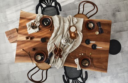 Overhead view of a wooden dining table with a beige cloth, dishes and bowls, a teapot, cutlery, and napkins, surrounded by assorted chairs.