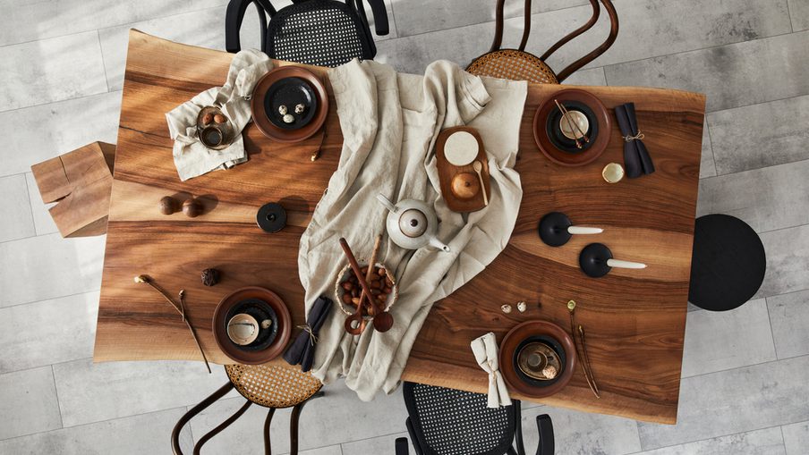 Overhead view of a wooden dining table with a beige cloth, dishes and bowls, a teapot, cutlery, and napkins, surrounded by assorted chairs.