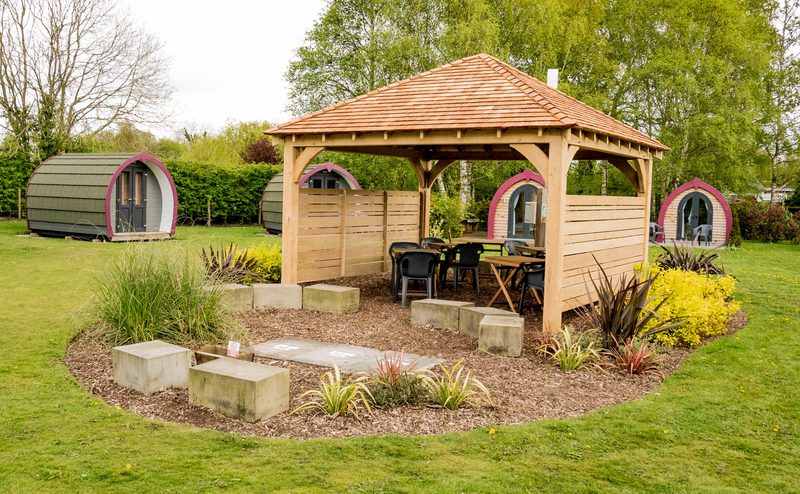 Three rounded pod cabins with purple trim surround a wooden gazebo in a green garden, with seating under the gazebo and a circular mulch bed nearby.