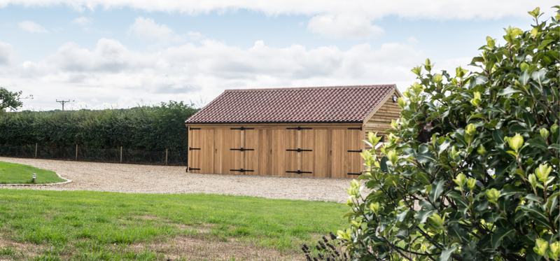 A long wooden shed with a red-tiled roof sits on a curved gravel driveway, bordered by green grass and a hedge, with a leafy shrub in the foreground.