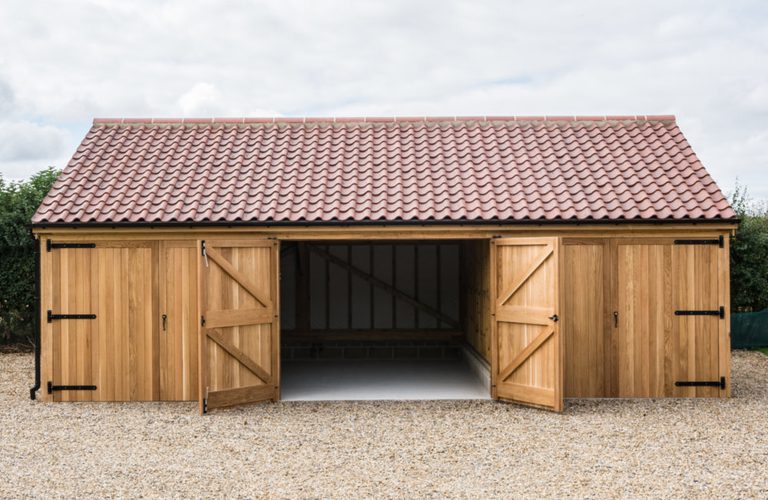 Wooden garden shed with a red-tiled roof and three bays; center bay open showing interior framing, outer bays closed, gravel ground.