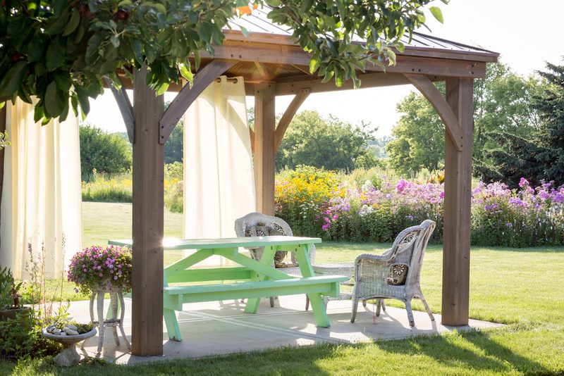 Wood pergola with cream curtains, a mint-green picnic table, and wicker chairs on a sunny patio, surrounded by blooming garden flowers.