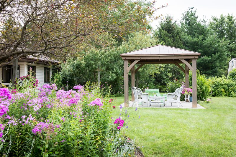 Backyard garden with a wooden gazebo and white outdoor seating on a concrete pad, surrounded by pink flowers, a green lawn, and trees.