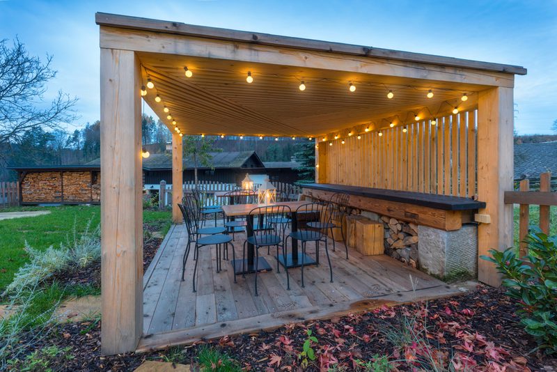 Wooden pergola patio with string lights, metal chairs around a table, a long bar with stacked firewood, and a garden yard at dusk.