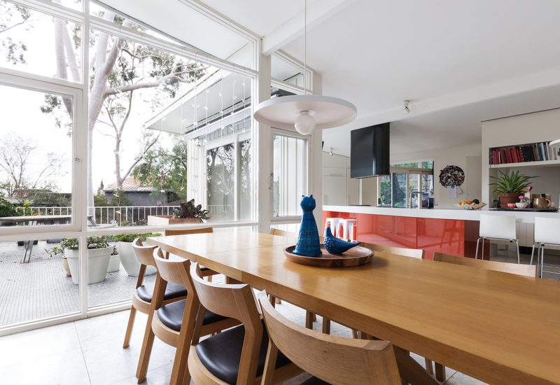 Bright open-plan kitchen and dining area with a long wooden table, matching chairs, large windows, red island, and blue ceramic figurines on a tray.