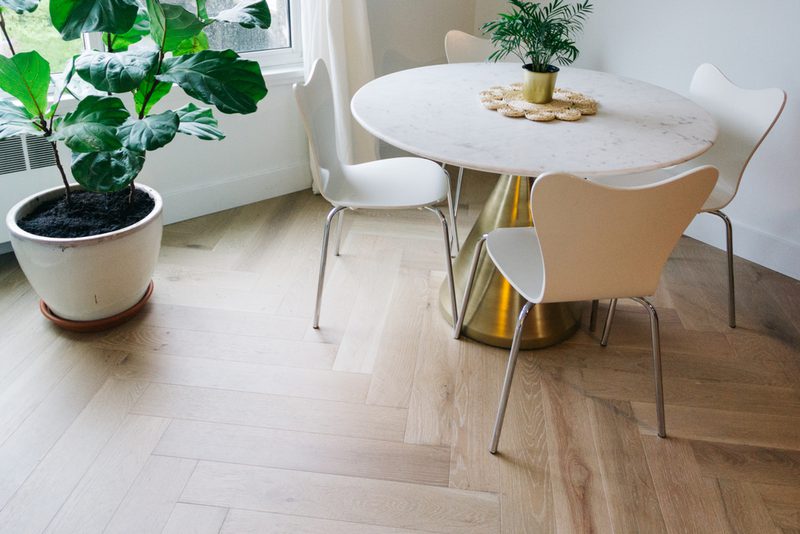 Bright dining area with a round marble table on a gold base, white chairs, a small plant centerpiece, and a large potted plant by the window; light wood floor.