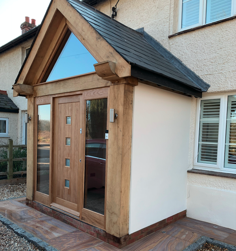 Wood-framed front porch with a gabled slate roof on a white house; glass side panels flank a paneled wooden door, with a triangular window above.