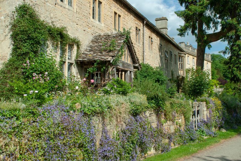 Stone house with ivy-covered walls and a wooden porch, bordered by a stone wall draped in purple flowers and lush garden foliage on a sunny day.