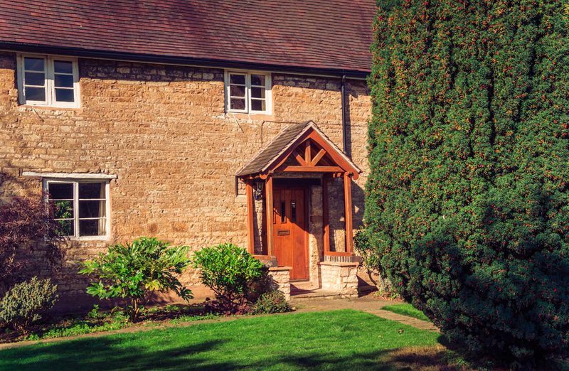 Stone cottage with a wooden gabled porch, brown door, red-tiled roof, and a green lawn; shrubs at the front and a tall evergreen hedge on the right.
