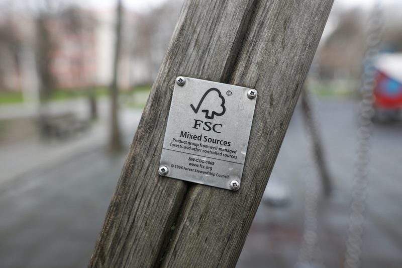 Close-up of a weathered wooden post with an FSC plaque displaying 'Mixed Sources' and logo; blurred park playground in the background.