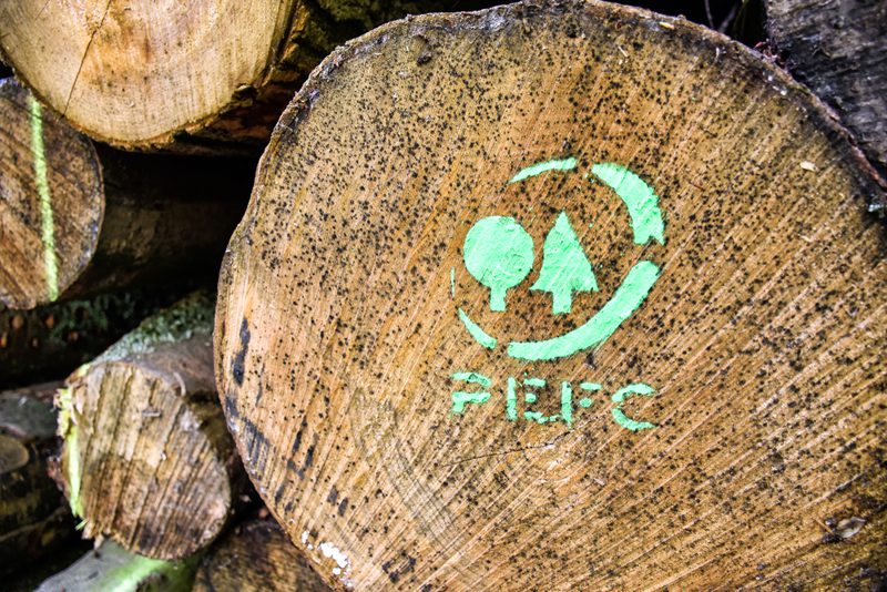 Close-up of a log cross-section with a green PEFC certification logo featuring trees; background shows stacked logs.
