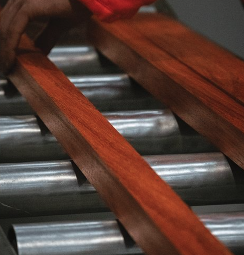 Close-up of reddish-brown wooden slats resting on a metal roller conveyor, with a gloved hand visible in the upper left.