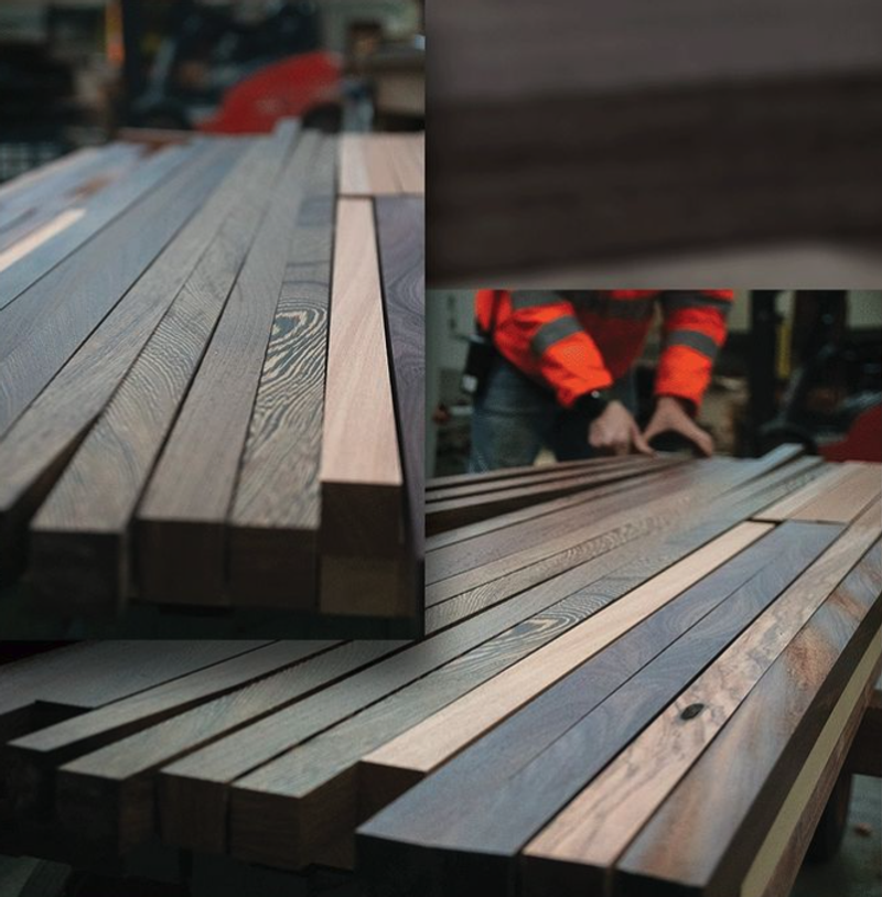 Stacks of wooden planks on a workshop bench, with a worker in a red jacket handling boards in the background.