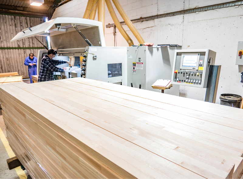 Woodworking shop with a long stack of pale planks in the foreground; a large industrial machine with a control panel and two workers nearby under yellow dust hoses.