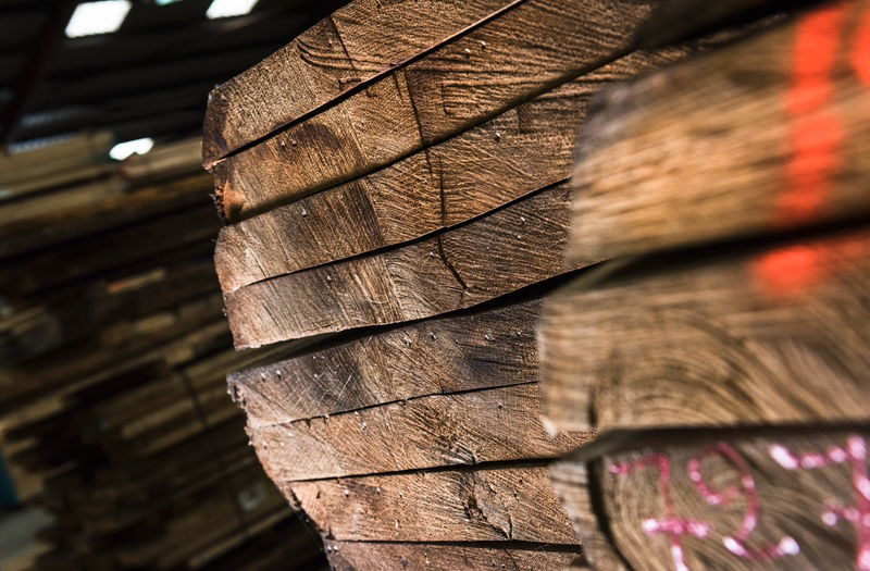 Close-up of rough-cut timber planks stacked in a lumberyard; visible grain and growth rings, nails along the edges, with other stacks blurred in the background.