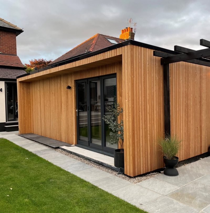 A modern wooden extension with vertical timber cladding and black-framed glass doors, flanked by a paved path, potted plants, lawn, and a black pergola.