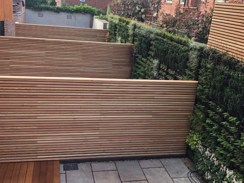 Outdoor courtyard with layered horizontal wooden slat privacy fences, green hedges along the right, and a gray stone tile patio in the foreground.