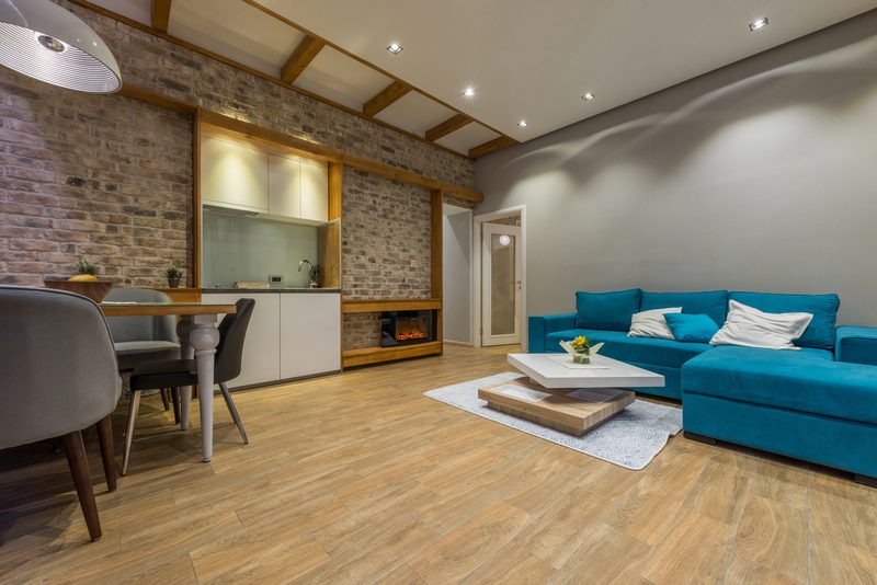 Spacious open-plan lounge with exposed brick walls, wooden ceiling beams, and a blue sectional sofa. A white square coffee table sits on a light rug beside a compact kitchen.