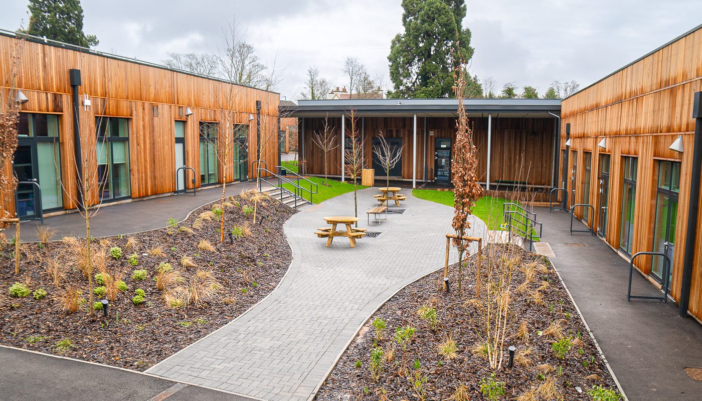Modern school courtyard with curved pathways, wooden cladding, landscaped gardens, and picnic tables for outdoor learning and relaxation.