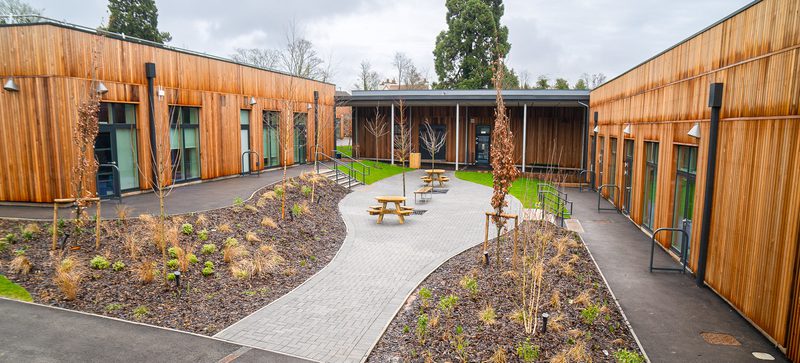 Courtyard between wood-paneled buildings with a curved paved walkway, picnic tables, bare trees, and mulched plant beds under a cloudy sky.
