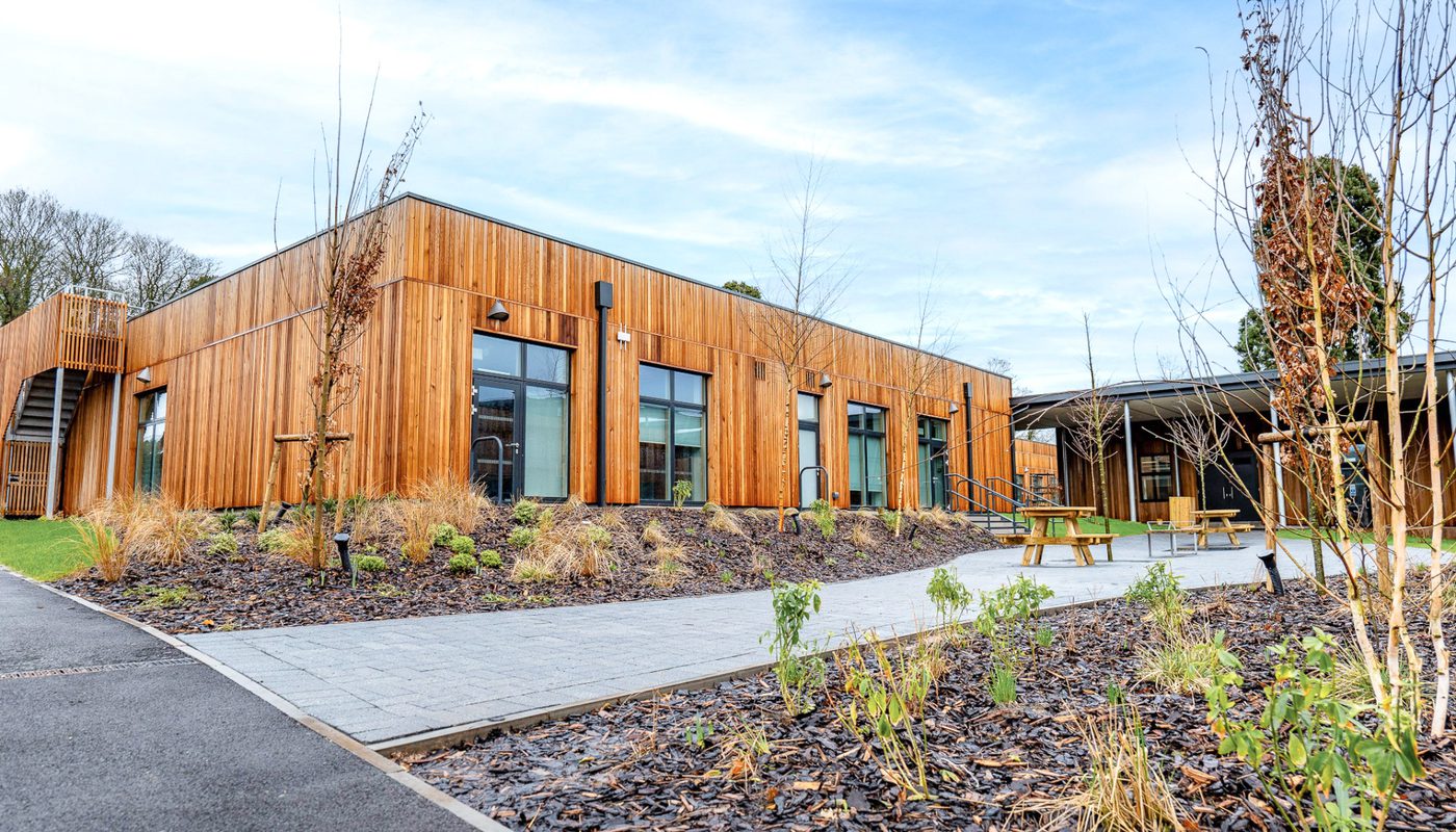 Eco-friendly school building with timber cladding, large windows, landscaped gardens, and outdoor seating areas for sustainable learning spaces.
