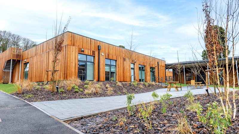 Modern wood-clad building with large glass doors, outdoor seating, and landscaped beds with young trees; paved paths in a campus courtyard.