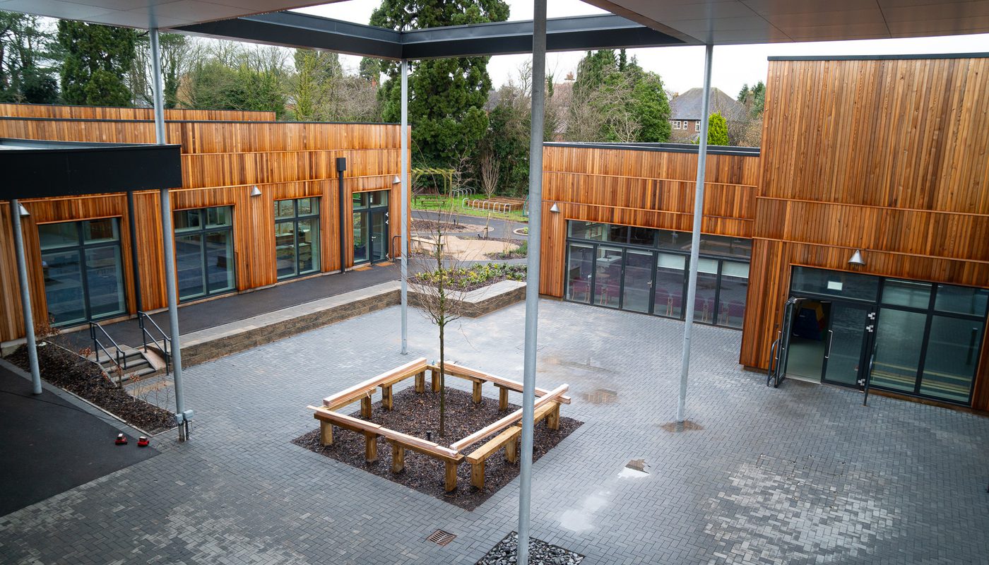 Spacious modern school courtyard with wooden cladding, large glass doors, a central tree planter, and covered walkways for outdoor learning and recreation.