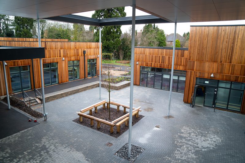 Open courtyard with timber-clad buildings, glass doors, and metal columns; a central planter with a small tree encircled by a wooden bench.