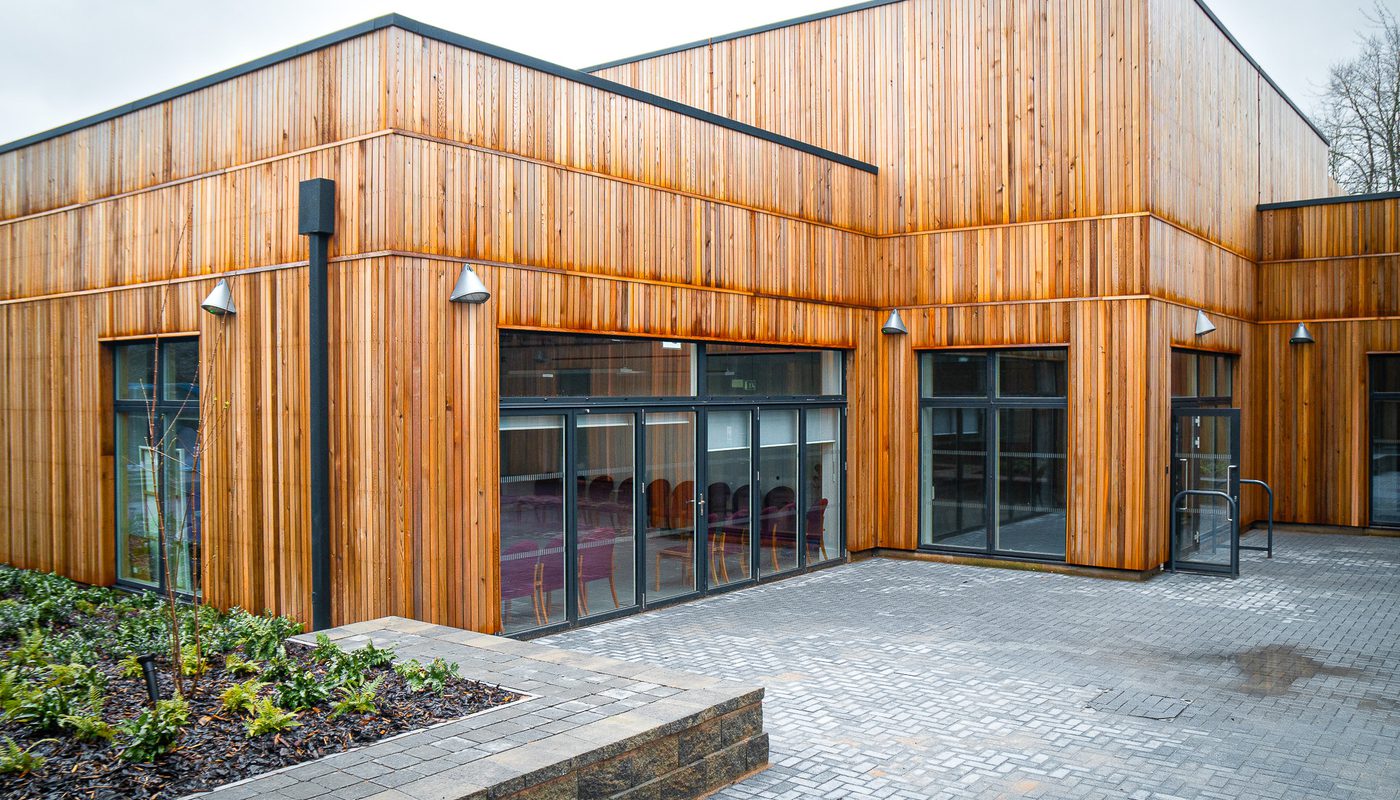 Modern school building with vertical timber cladding, large glass windows, and a paved courtyard with landscaped greenery.