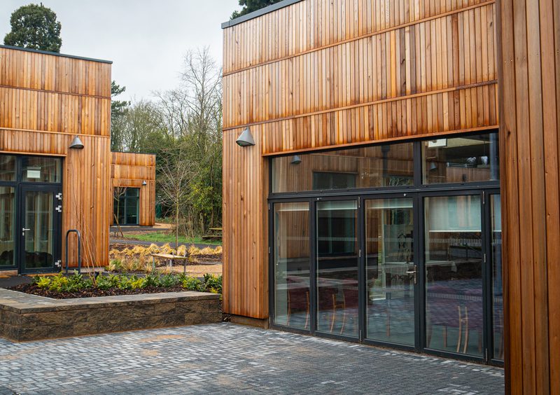 Modern wooden-clad building with large glass doors and black metal frames, a stone planter and cobblestone courtyard, with trees in the background.