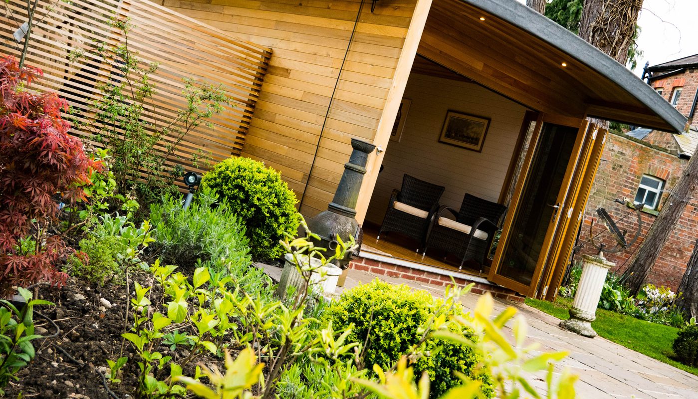 Wood-clad garden studio with open glass doors, two wicker chairs inside, a privacy screen, and a lush foreground of shrubs and a stone path.