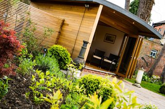 Wood-clad garden studio with open glass doors, two wicker chairs inside, a privacy screen, and a lush foreground of shrubs and a stone path.