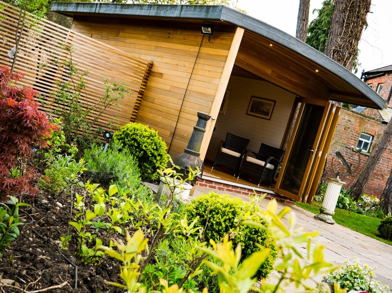 Wood-clad garden studio with open glass doors, two wicker chairs inside, a privacy screen, and a lush foreground of shrubs and a stone path.