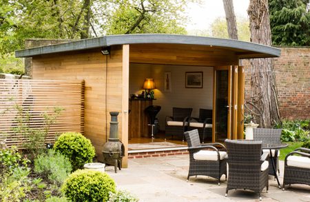 Wooden garden studio with a curved roof opens to a patio; wicker seating and a small table outside, plus a chiminea, lush plants, and a brick wall in the background.