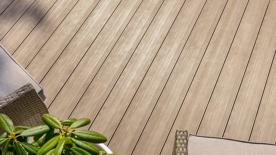 Top-down view of a sunlit wooden deck with diagonal planks, a wicker lounge chair with a light cushion, and a green plant in the bottom left.
