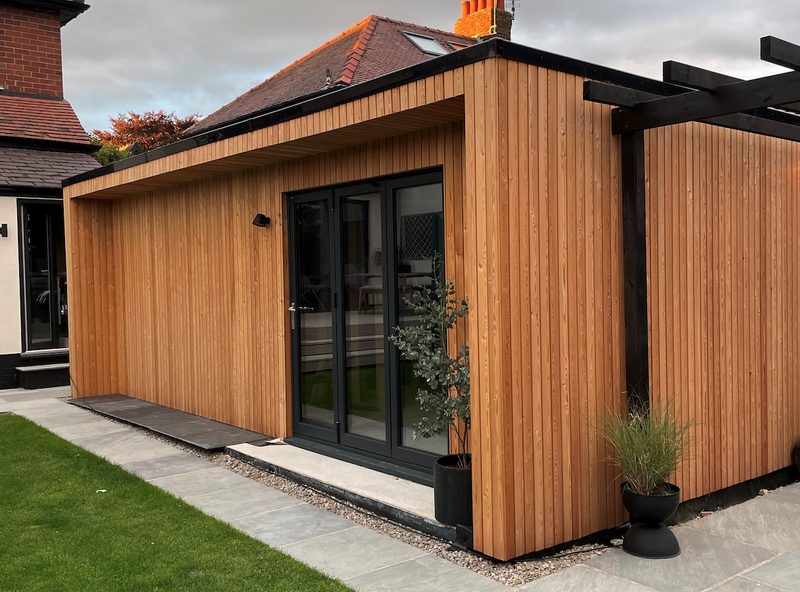 Modern wooden garden room with vertical slats, black-framed glass doors, a wall light, and potted plants beside a gravel path and green lawn.