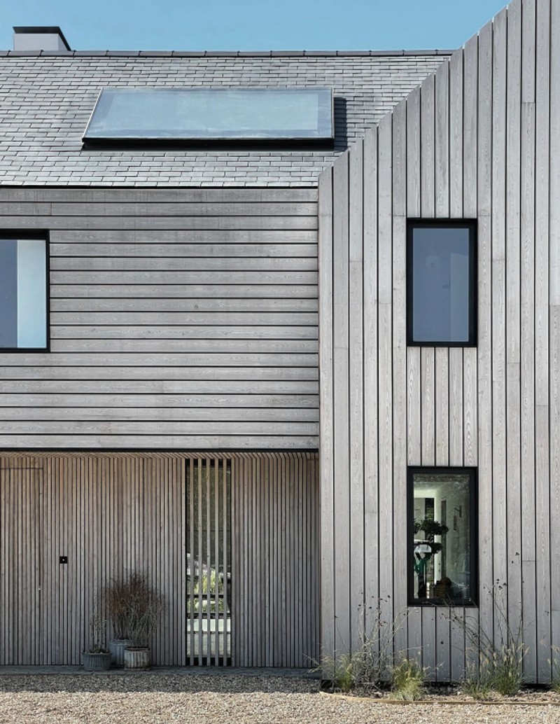 Modern gray wooden house with a sloped roof and skylight. Vertical siding on the right, a tall narrow window, and a slatted entry with potted plants.