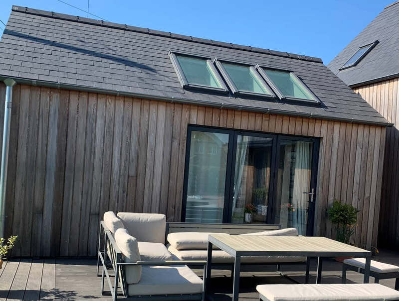 Wood-clad house with a sloped roof and skylights, glass sliding doors, and a beige outdoor sofa set with a rectangular table on a wooden deck.
