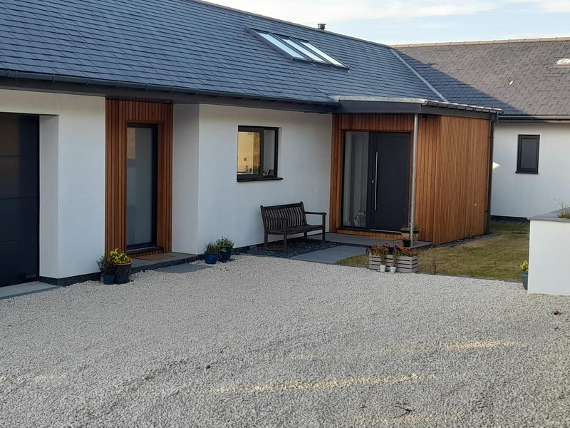 Modern, single-story house with white walls and vertical wooden cladding; gravel driveway, bench, potted plants, and a small lawn.