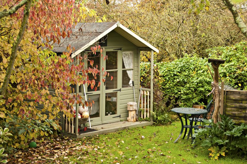 Green wooden garden shed with glass doors and a small porch, surrounded by autumn leaves; a round metal table and chairs sit on the lawn to the right.