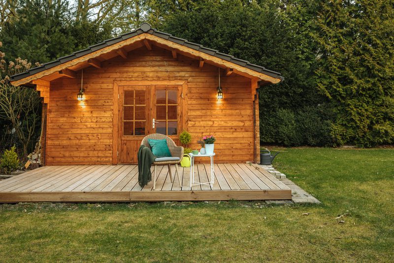 Wooden cabin on a raised deck in a garden, with a wicker chair, green blanket, small white table, potted plants, and hanging lantern lights.