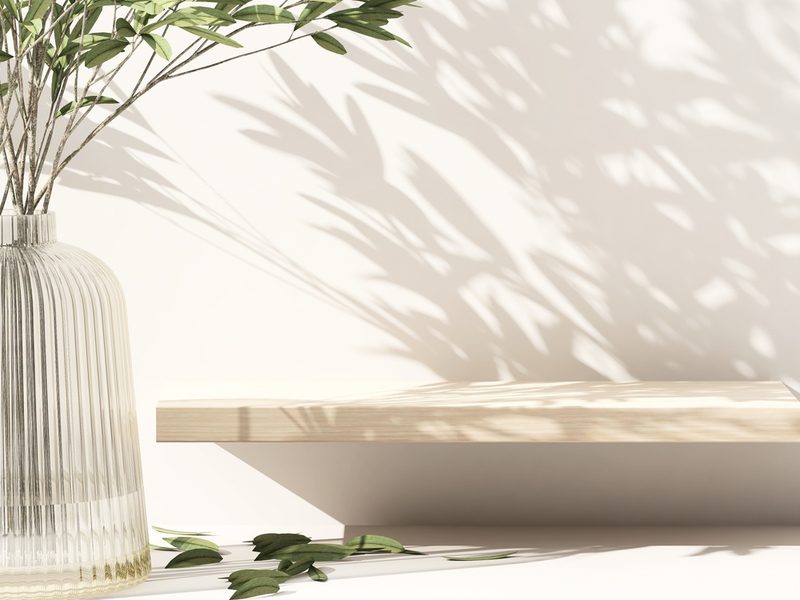 Tall, clear ribbed glass vase with green stems sits on a light surface beside a light wooden shelf; leaf shadows play across the pale wall.