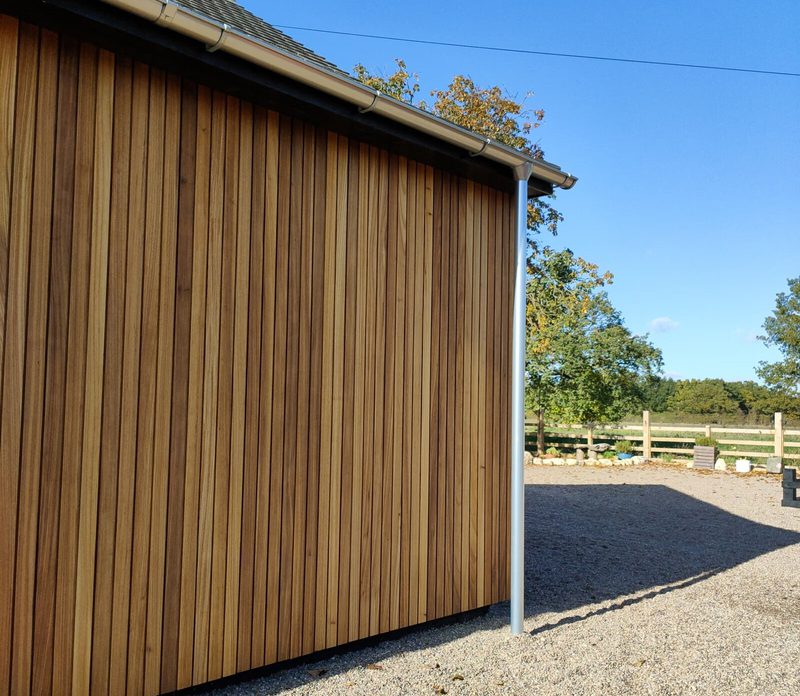 Wooden shed with vertical brown planks, white gutter and downspout, gravel yard, and a rural fence with trees under a blue sky.