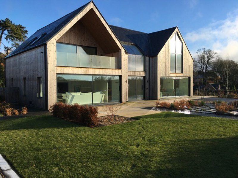 Modern wooden house with large glass walls and a glass balcony, featuring A-frame roof sections and a green lawn under a clear blue sky.