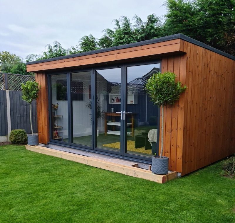 A wooden garden room with expansive glass doors, two potted topiary trees, and a green lawn in front.