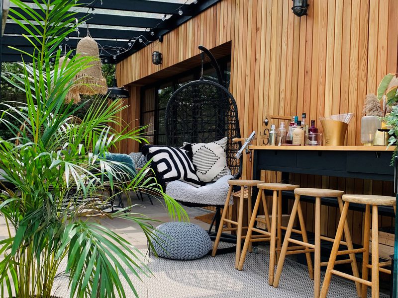 Outdoor patio with vertical wooden siding, string lights, and a black wicker hanging chair with geometric cushions; a bar with stools and bottles beside lush plants.