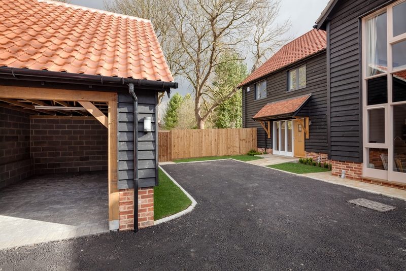 Two dark wooden houses with red-tiled roofs flank a curved asphalt driveway, with a brick carport on the left and a wooden fence behind.