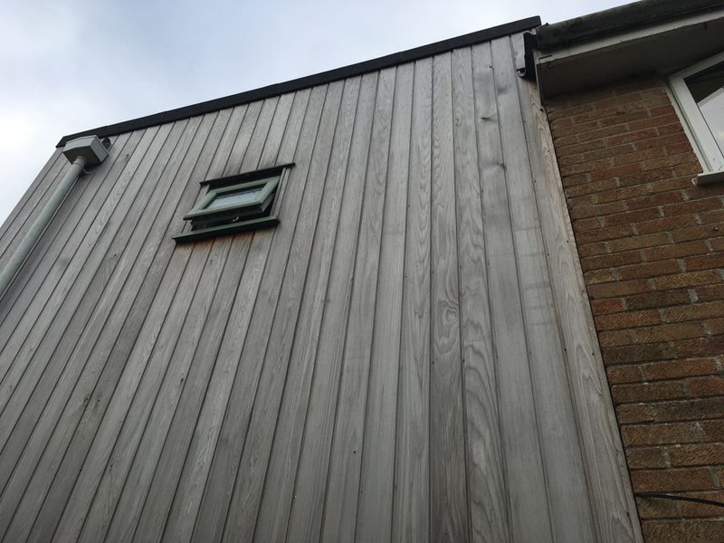 Side of a building with vertical gray weathered wooden siding, a small green-framed window, a left gutter, and a brick wall on the right under a pale sky.