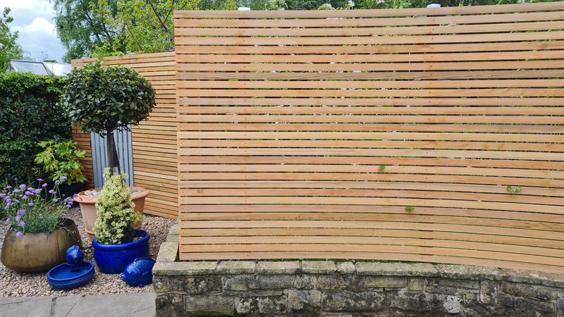 Garden scene featuring a horizontal wooden slat privacy screen, a stone base, potted topiary, blue pots, purple flowers, and gravel ground.